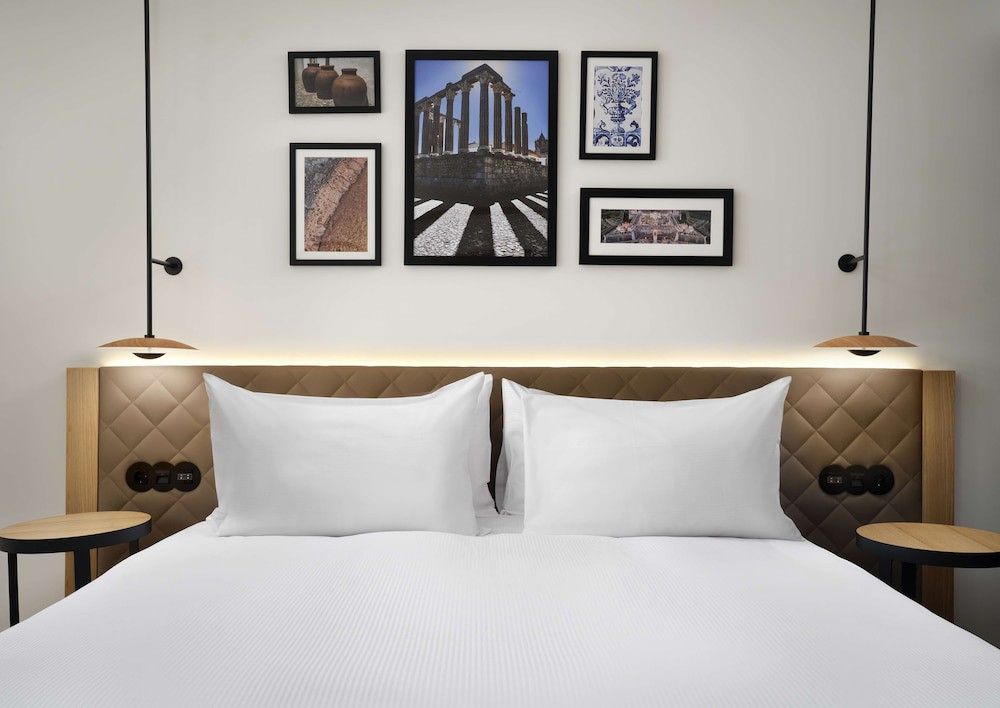Ett modernt hotellrum med en prydligt bäddad säng, vadderad sänggavel, två hängande lampor och en gallerivägg med inramad konst som föreställer romerska ruiner och keramik.