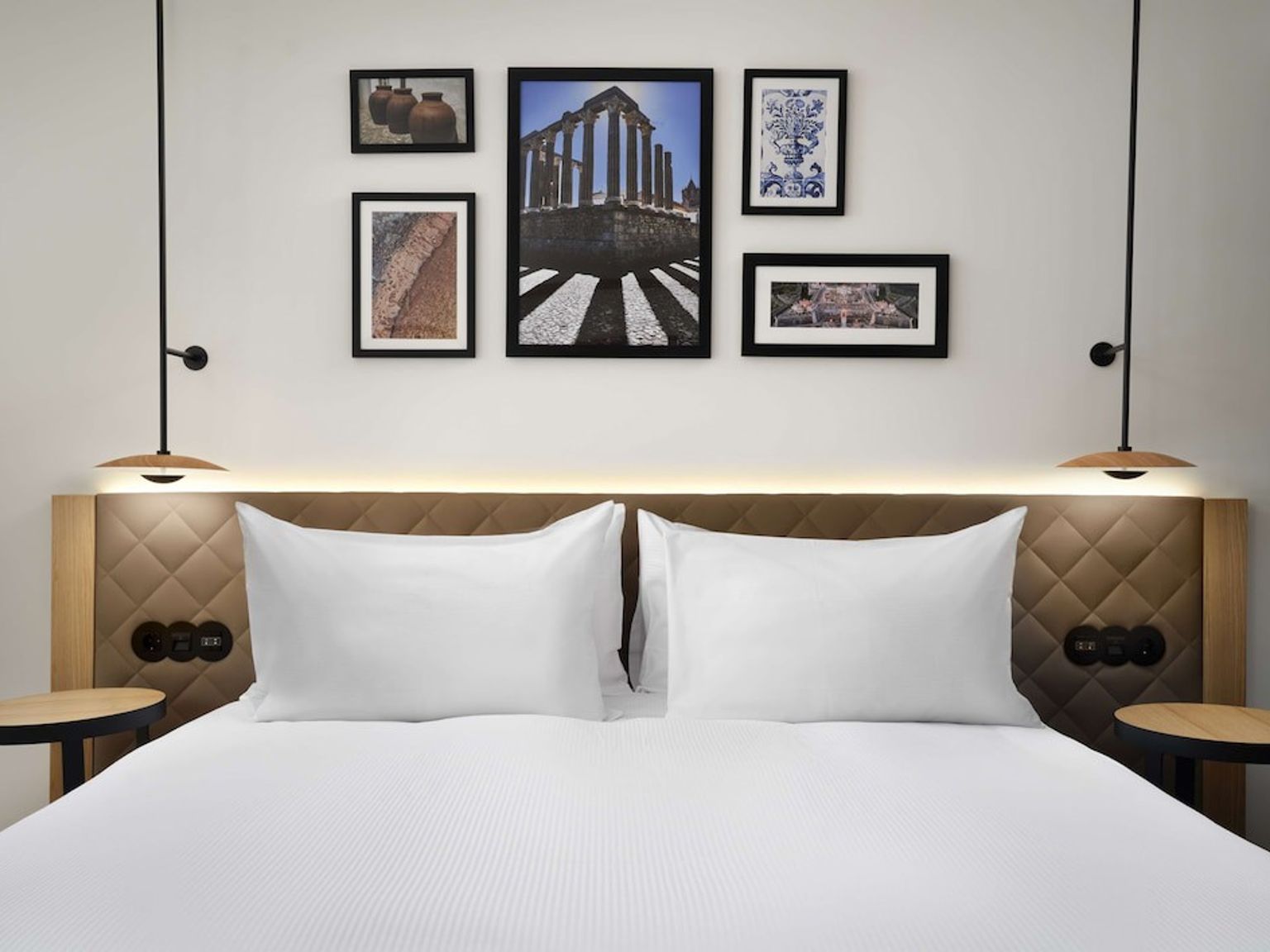 Ett modernt hotellrum med en prydligt bäddad säng, vadderad sänggavel, två hängande lampor och en gallerivägg med inramad konst som föreställer romerska ruiner och keramik.