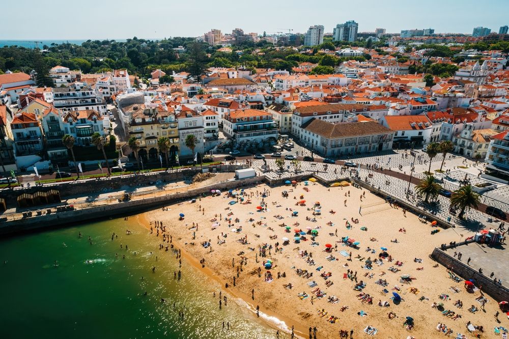 Flygfoto över en fullsatt strand intill en stad med byggnader och röda tegeltak.