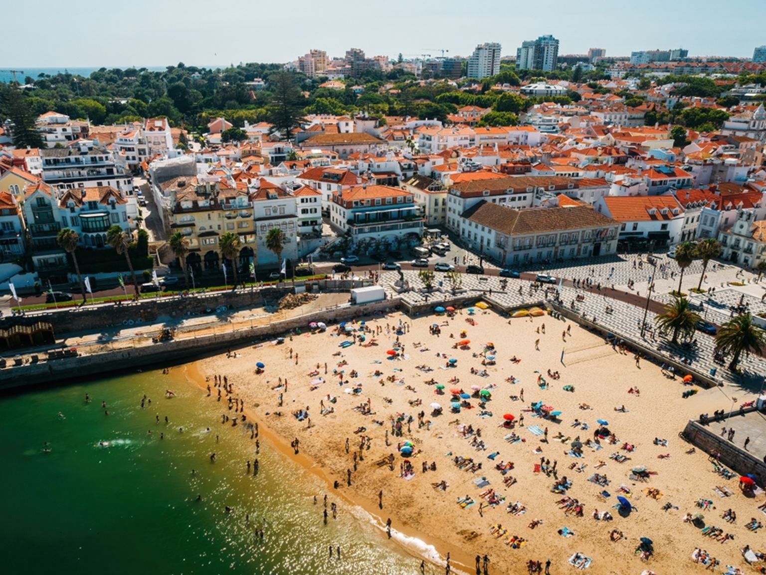 Flygfoto över en fullsatt strand intill en stad med byggnader och röda tegeltak.
