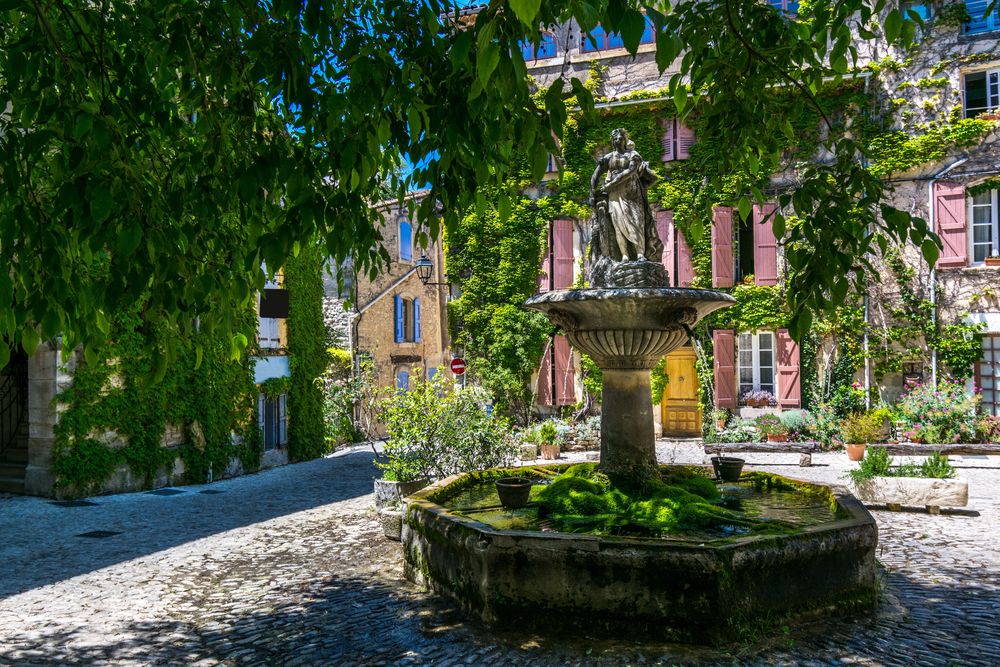 A stone fountain with a woman statue in a sunny, ivy-covered cobblestone village square with buildings featuring pink shutters. Saignon, Provence, France.