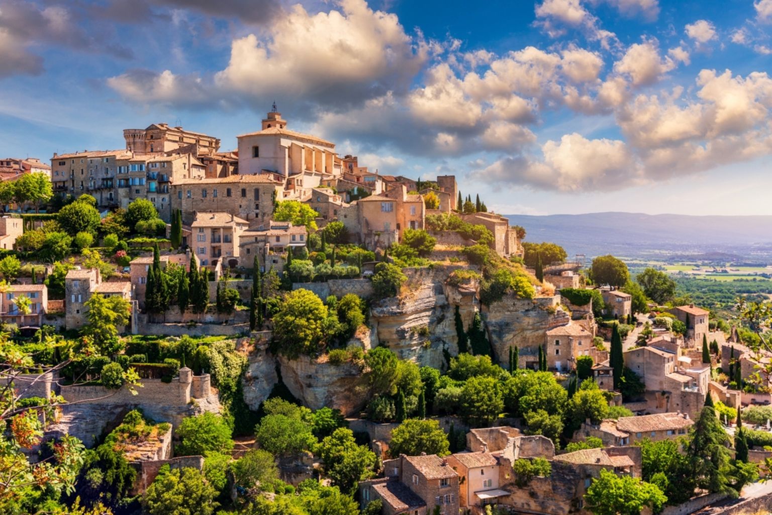 Picturesque hilltop village with ancient stone buildings, a prominent church, and lush trees under a blue sky with white clouds. Gordes, France