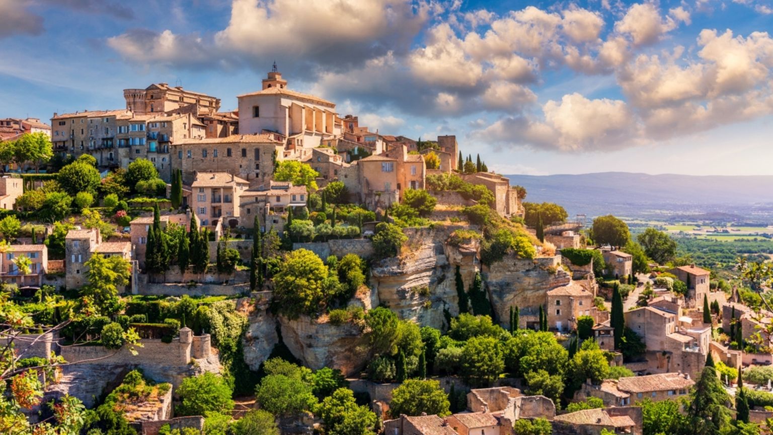 Picturesque hilltop village with ancient stone buildings, a prominent church, and lush trees under a blue sky with white clouds. Gordes, France
