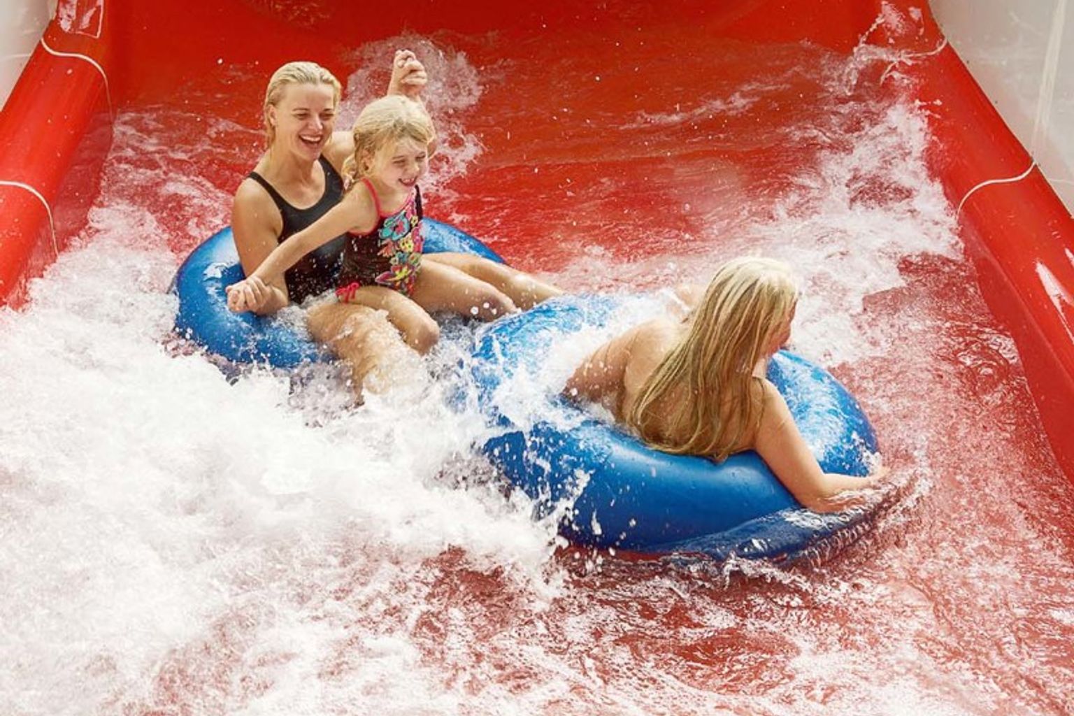 Två kvinnor och ett barn åker på blåa badringar nerför en röd vattenrutschkana, omgivna av stänk.