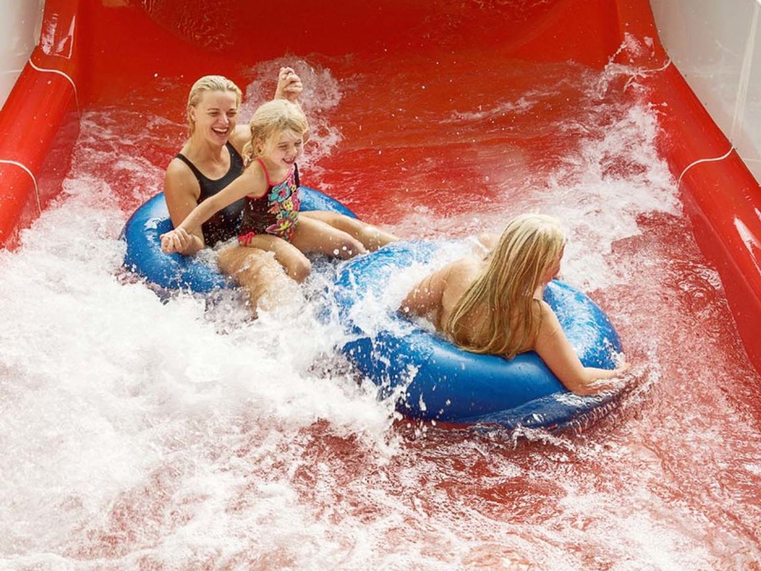 Två kvinnor och ett barn åker på blåa badringar nerför en röd vattenrutschkana, omgivna av stänk.