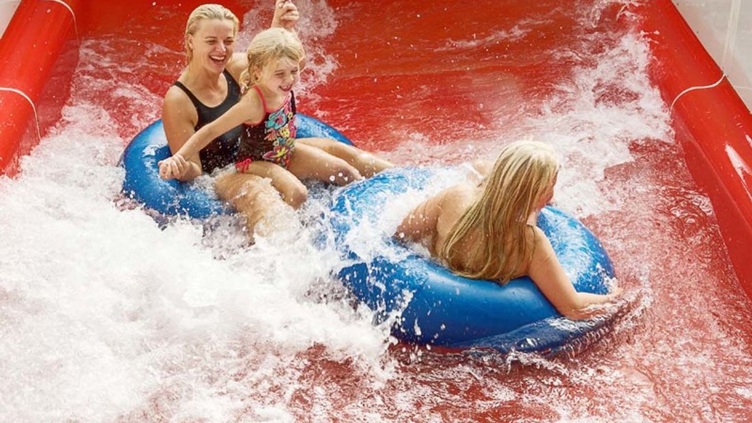 Två kvinnor och ett barn åker på blåa badringar nerför en röd vattenrutschkana, omgivna av stänk.