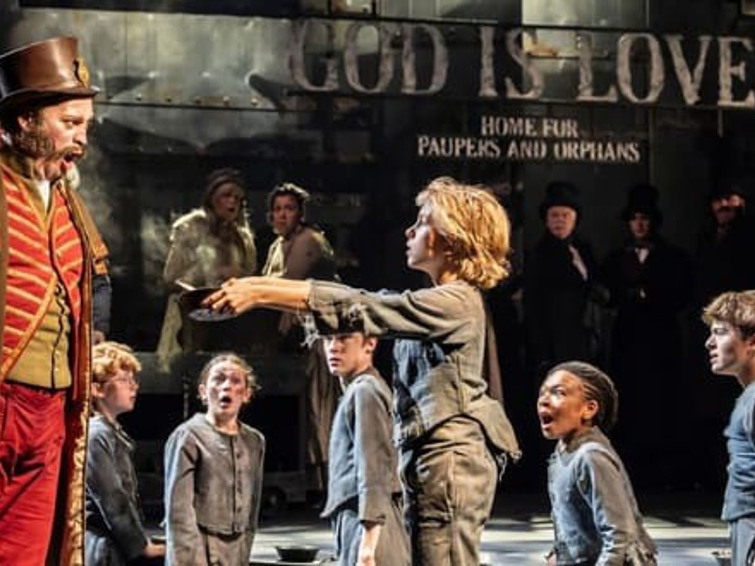 A stage scene shows a man in a period uniform shouting at a young boy holding an empty bowl, surrounded by other children and a sign reading "Home for Paupers and Orphans."