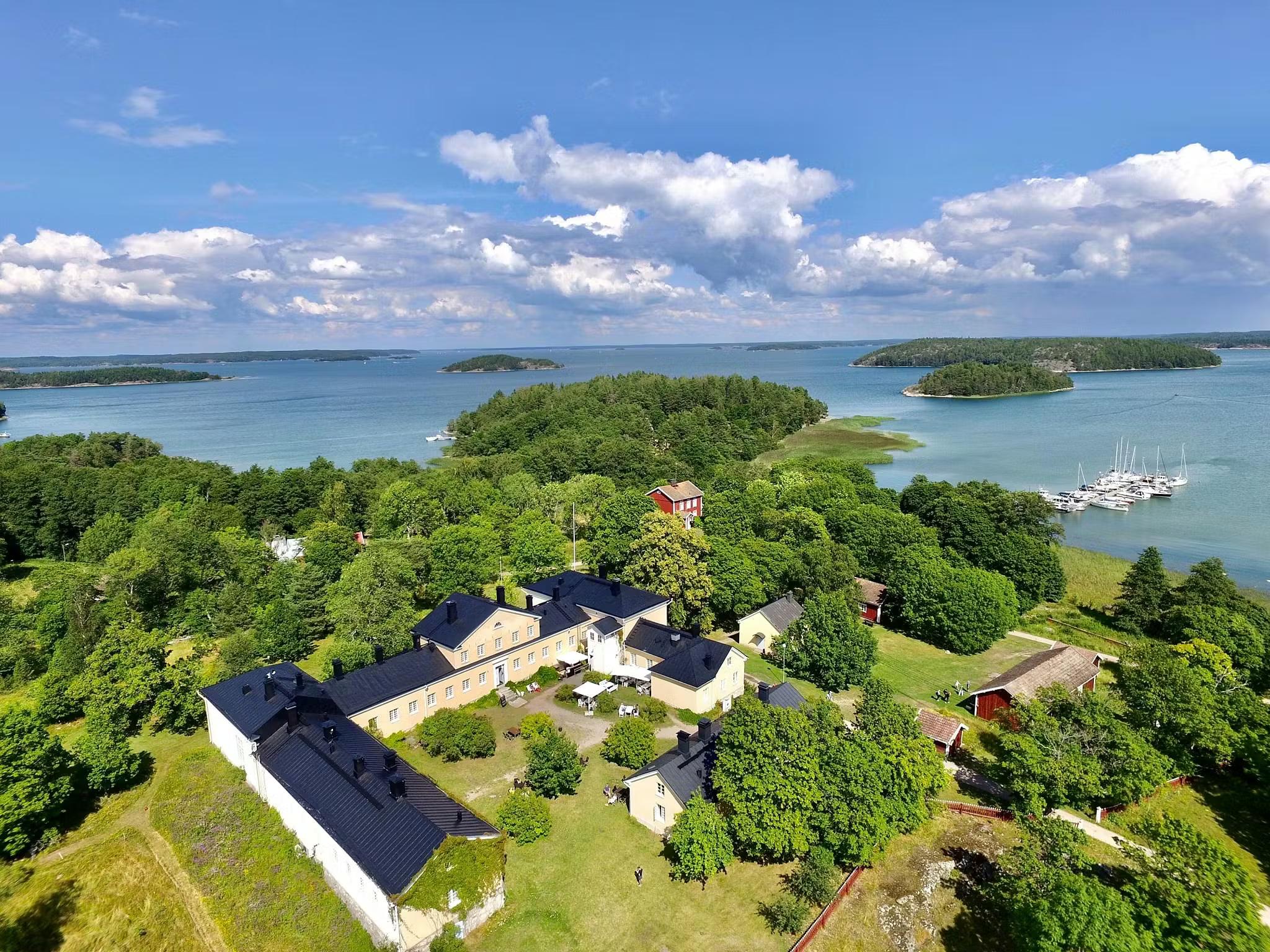 Aerial view of a coastal settlement with light-colored buildings and green trees, overlooking an archipelago with sailboats.