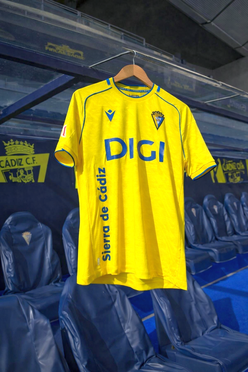 Żółta koszulka piłkarska z niebieskim napisem „DIGI” i herbem klubowym, wisząca w ławce rezerwowych na stadionie.
