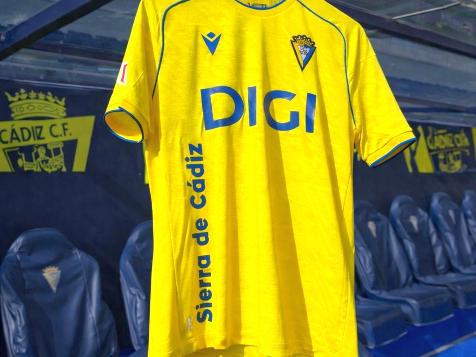 Uma camisa de futebol amarela com o texto azul "DIGI" e um brasão do clube, pendurada no banco de reservas de um estádio.