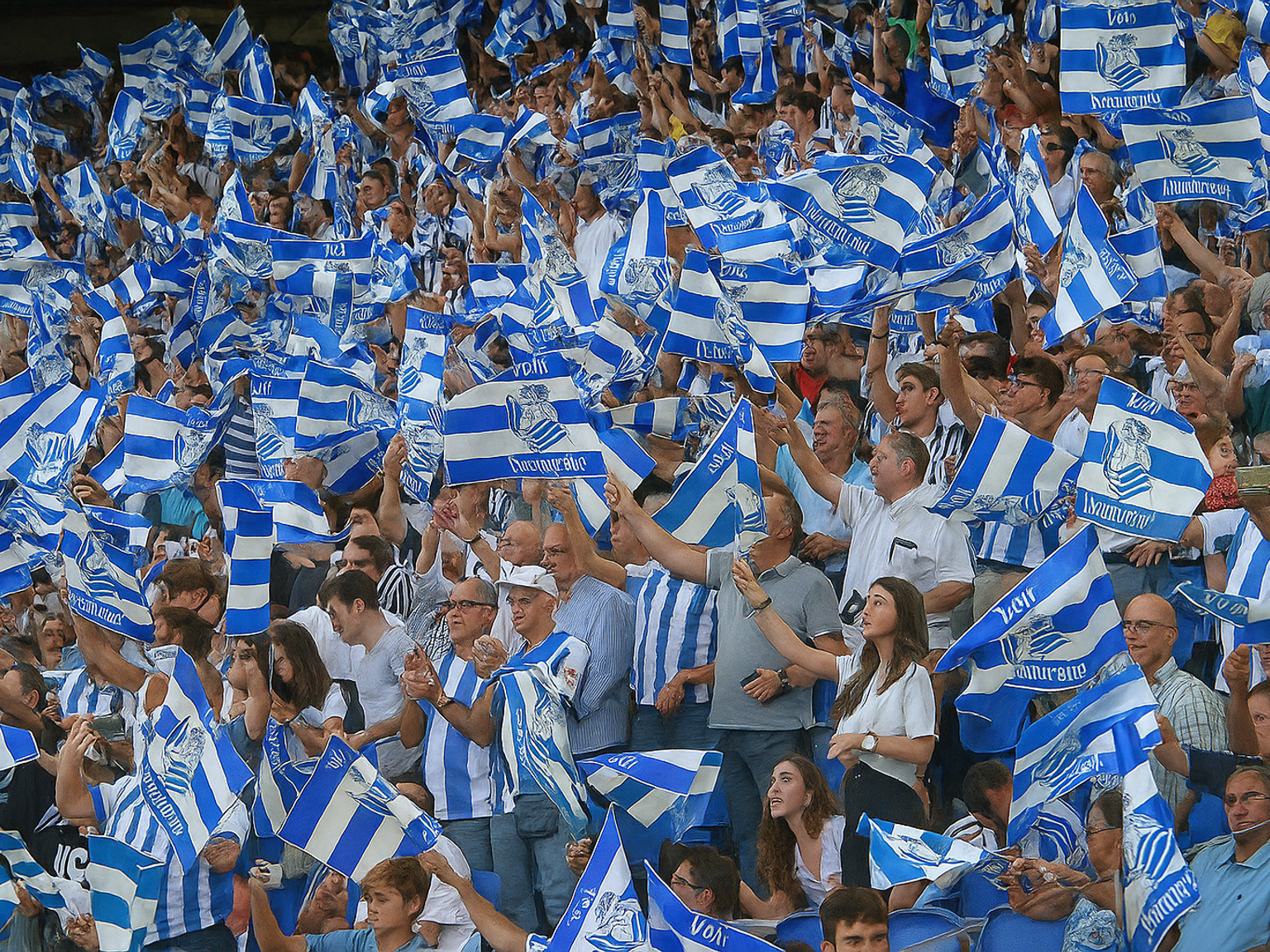 A large crowd of people waves numerous blue and white striped flags, many wearing matching attire.