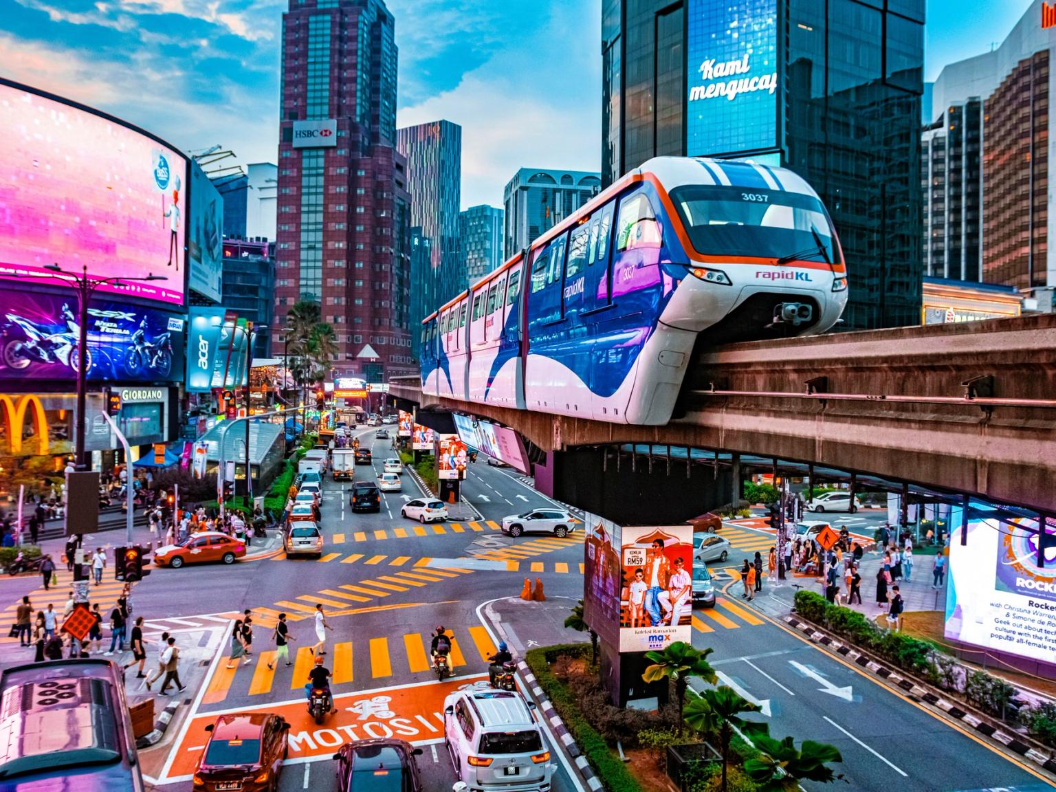 Travl bygadescene med en monorail ovenover, høje bygninger og lyse digitale reklametavler. Kuala Lumpur