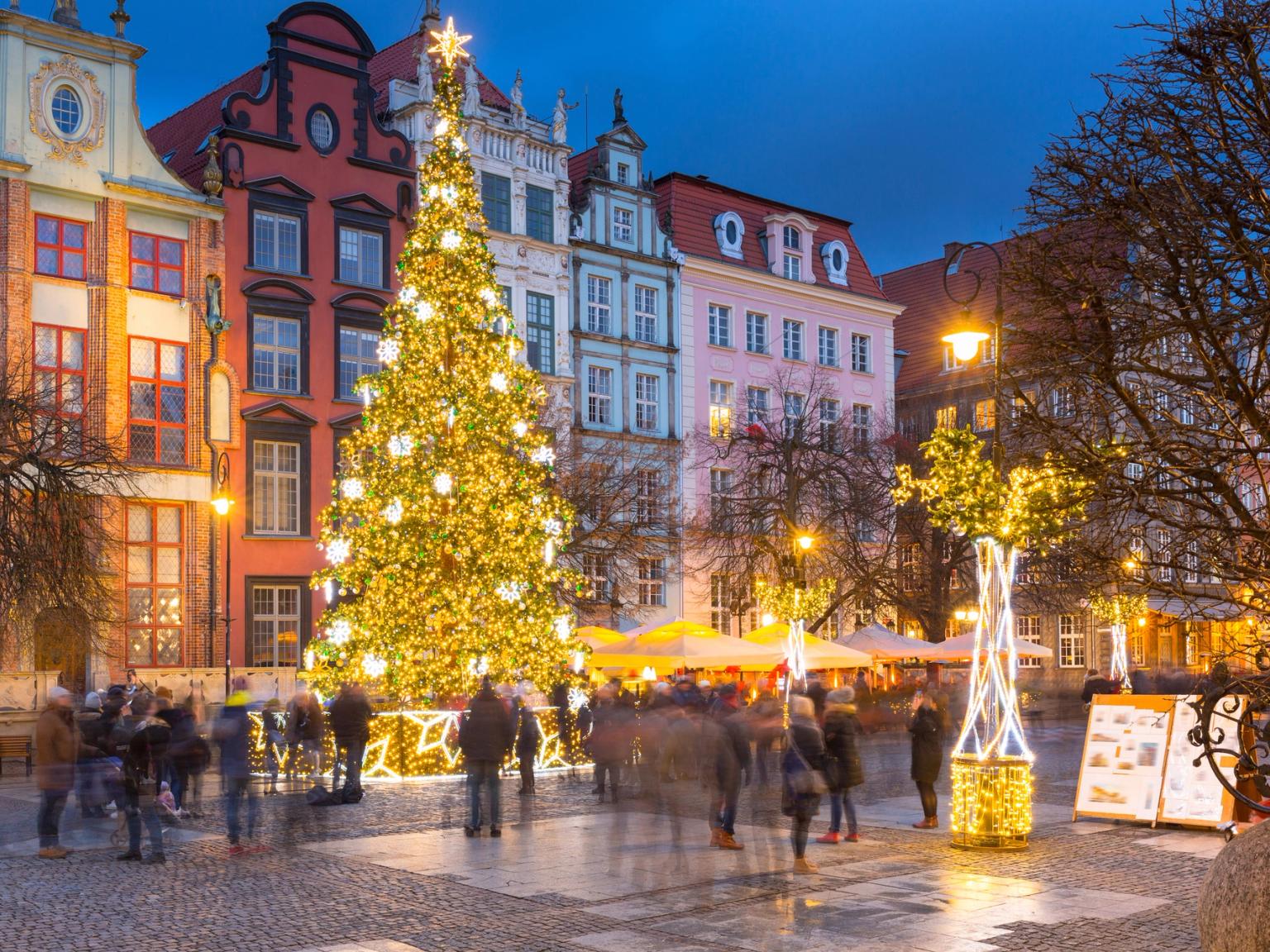 En livlig julmarknad i Gdansk i skymningen med en stor, upplyst julgran, folkmassor och historiska byggnader.