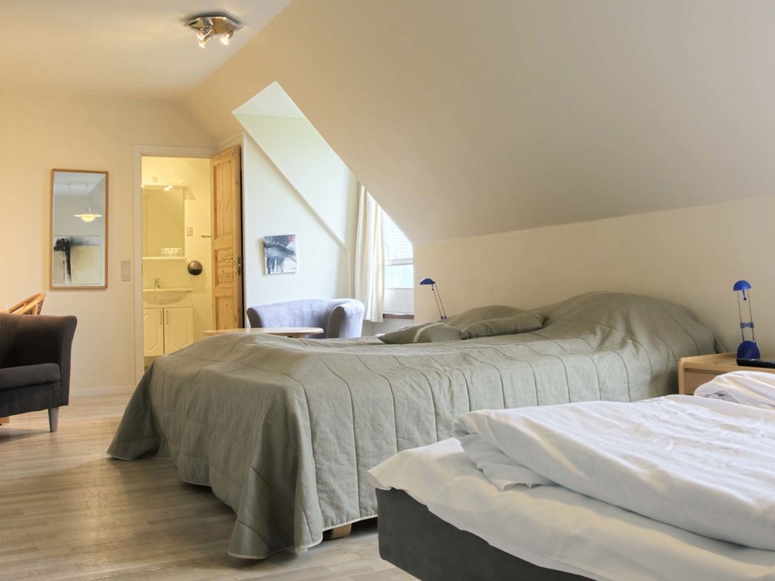 Ein heller Raum mit zwei Betten, einer schrägen Dachschräge, einem Sessel und einem Blick in ein Badezimmer. Hovborg Kro