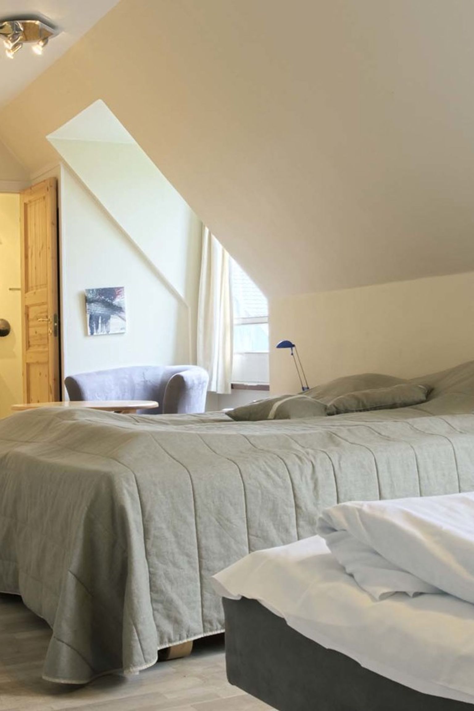 Ein heller Raum mit zwei Betten, einer schrägen Dachschräge, einem Sessel und einem Blick in ein Badezimmer. Hovborg Kro