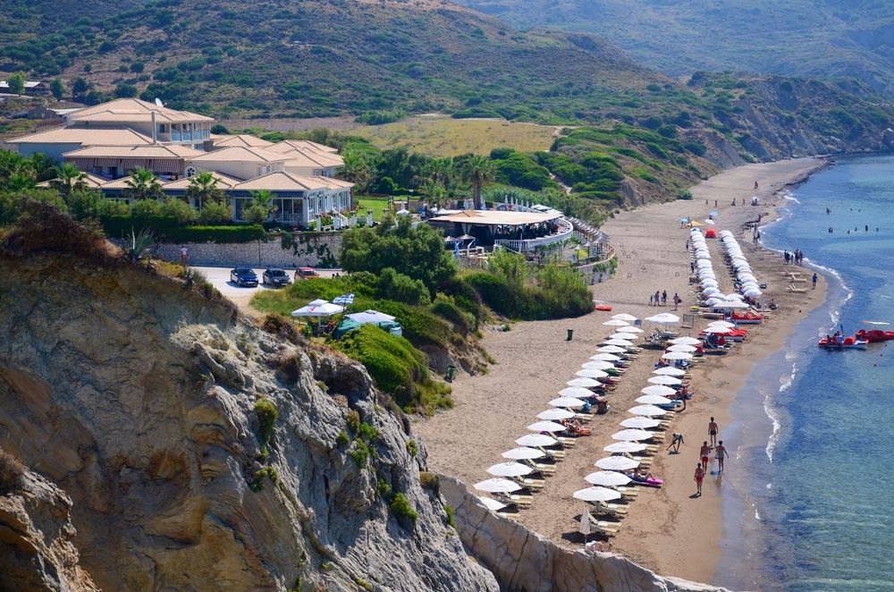 En resortbyggnad på en klippa med utsikt över en sandstrand kantad med vita parasoller och människor. Kalamaki, Zakhyntos