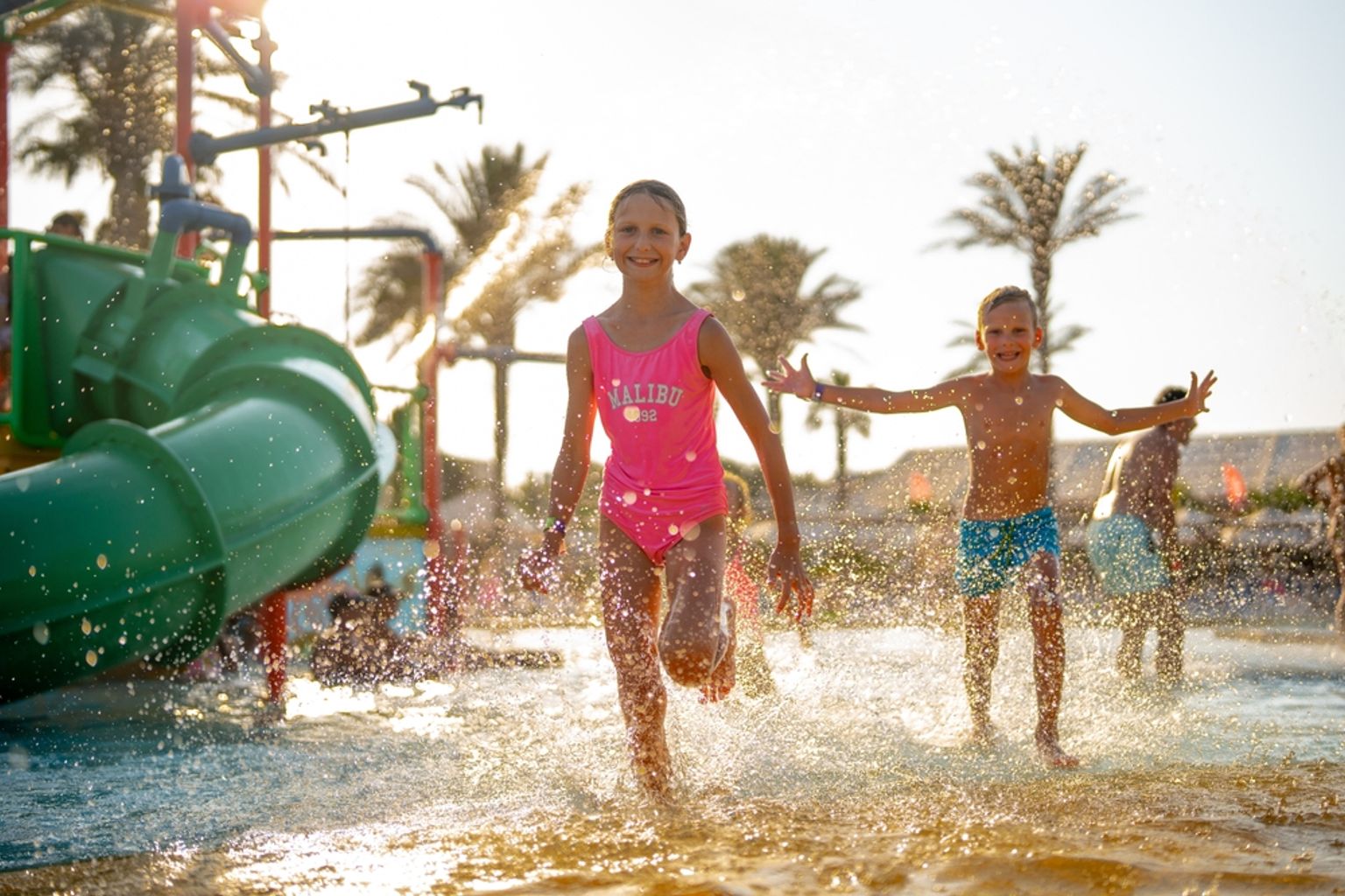 Två glada barn springer och plaskar i en solig vattenpark med en grön rutschkana.