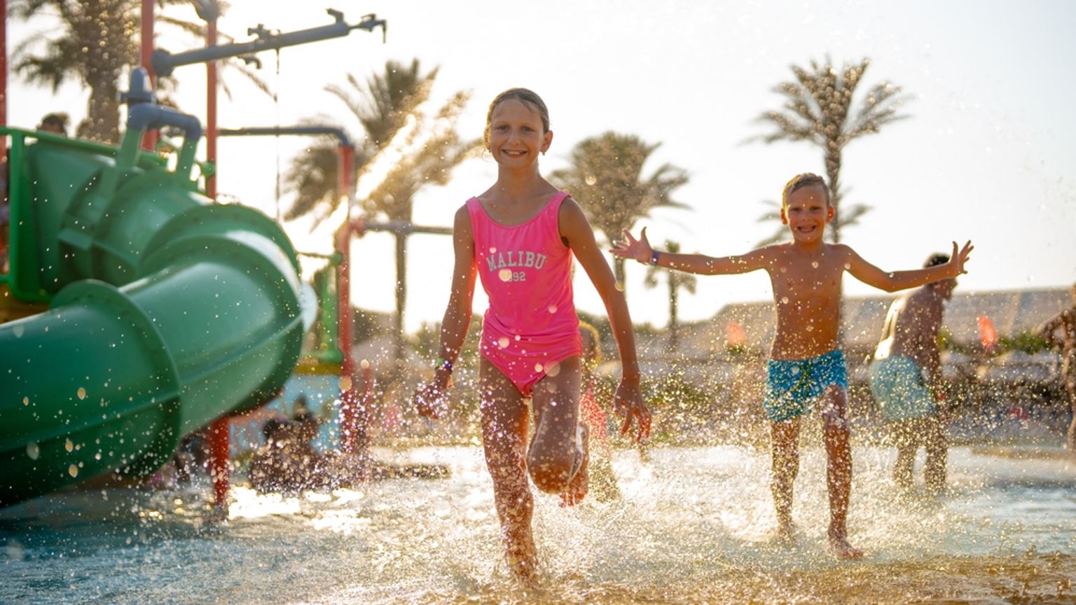 Två glada barn springer och plaskar i en solig vattenpark med en grön rutschkana.