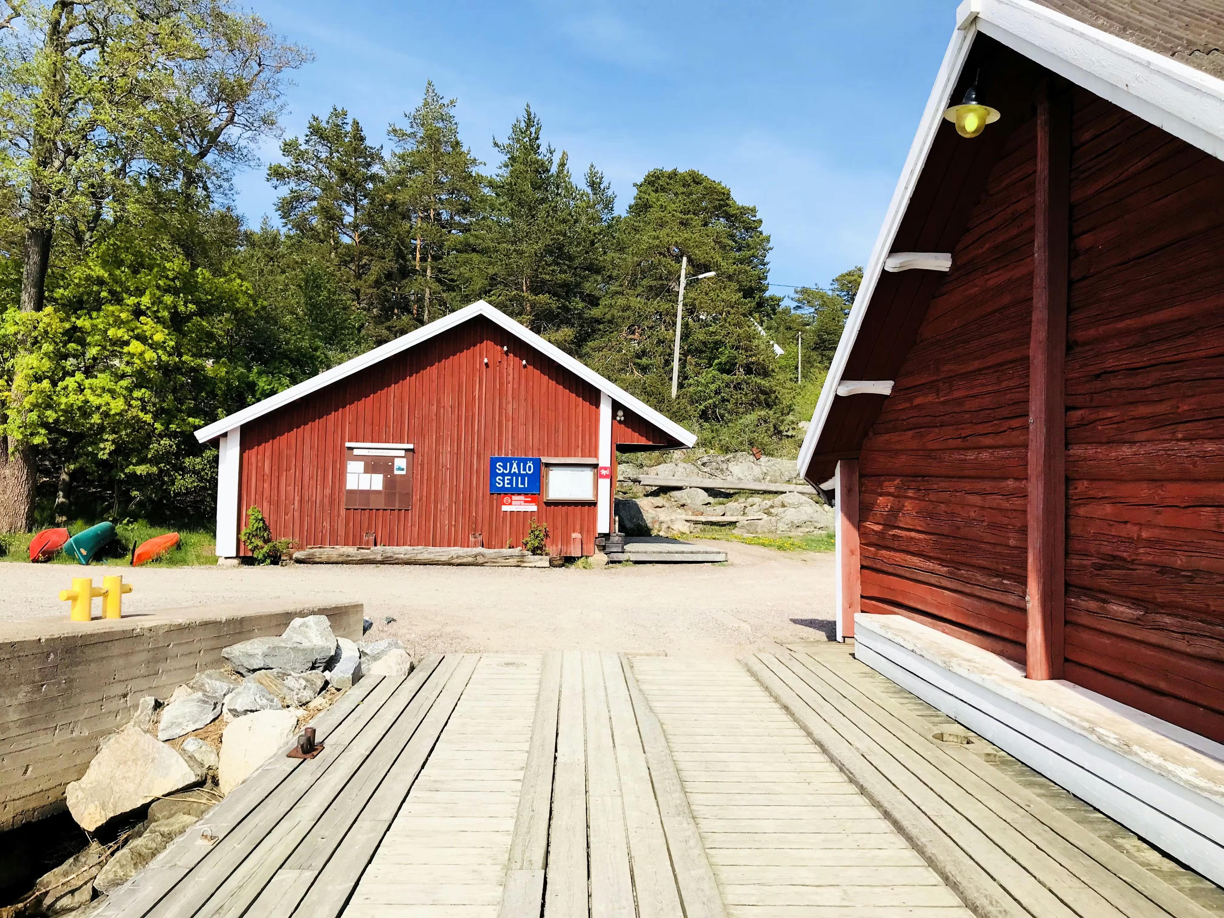 A wooden pier leads to a red building with a "SJÄLÖ SEILI" sign, next to another red building and green trees under a clear sky.