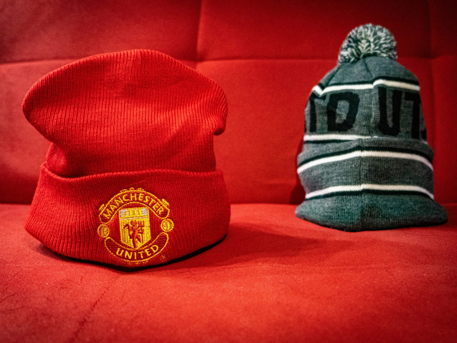 A red Manchester United beanie and a grey striped pom-pom hat on a red background.