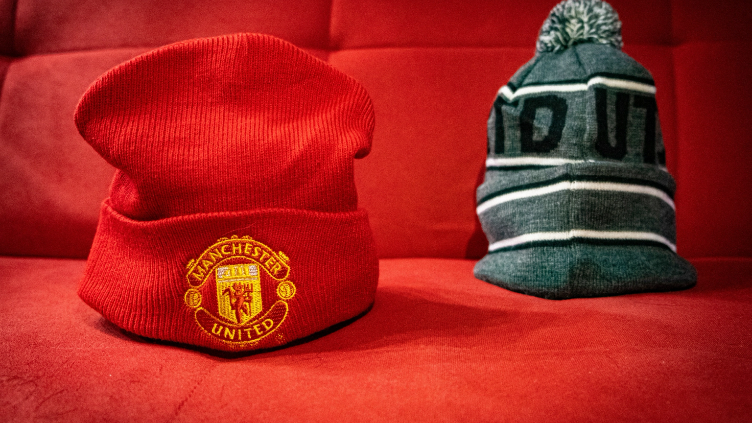 A red Manchester United beanie and a grey striped pom-pom hat on a red background.