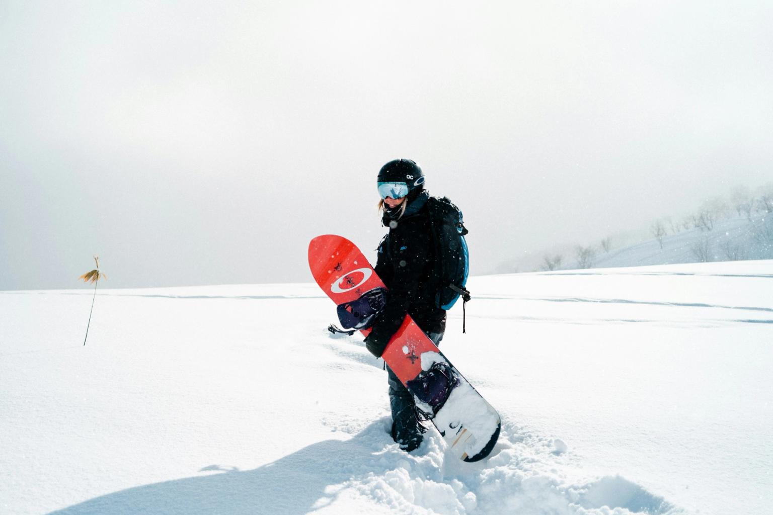 En snowboardkjører med hjelm og skibriller går gjennom dyp snø og bærer et rødt snowboard.