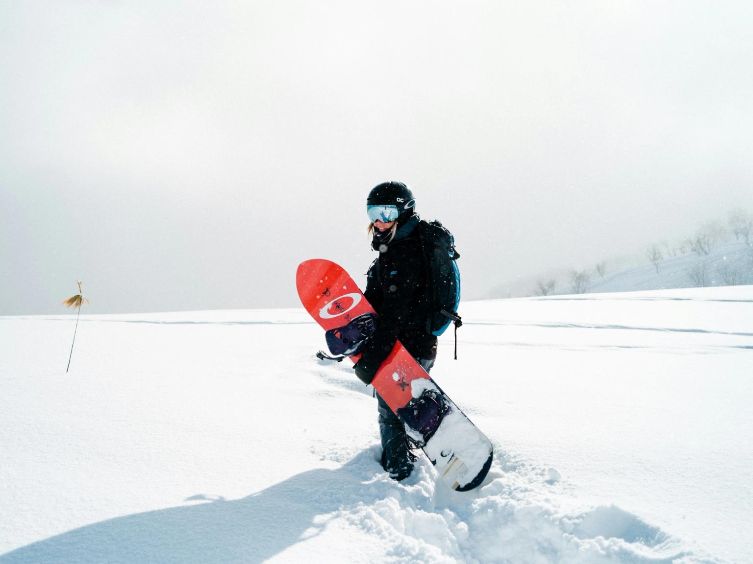 En snowboardkjører med hjelm og skibriller går gjennom dyp snø og bærer et rødt snowboard.