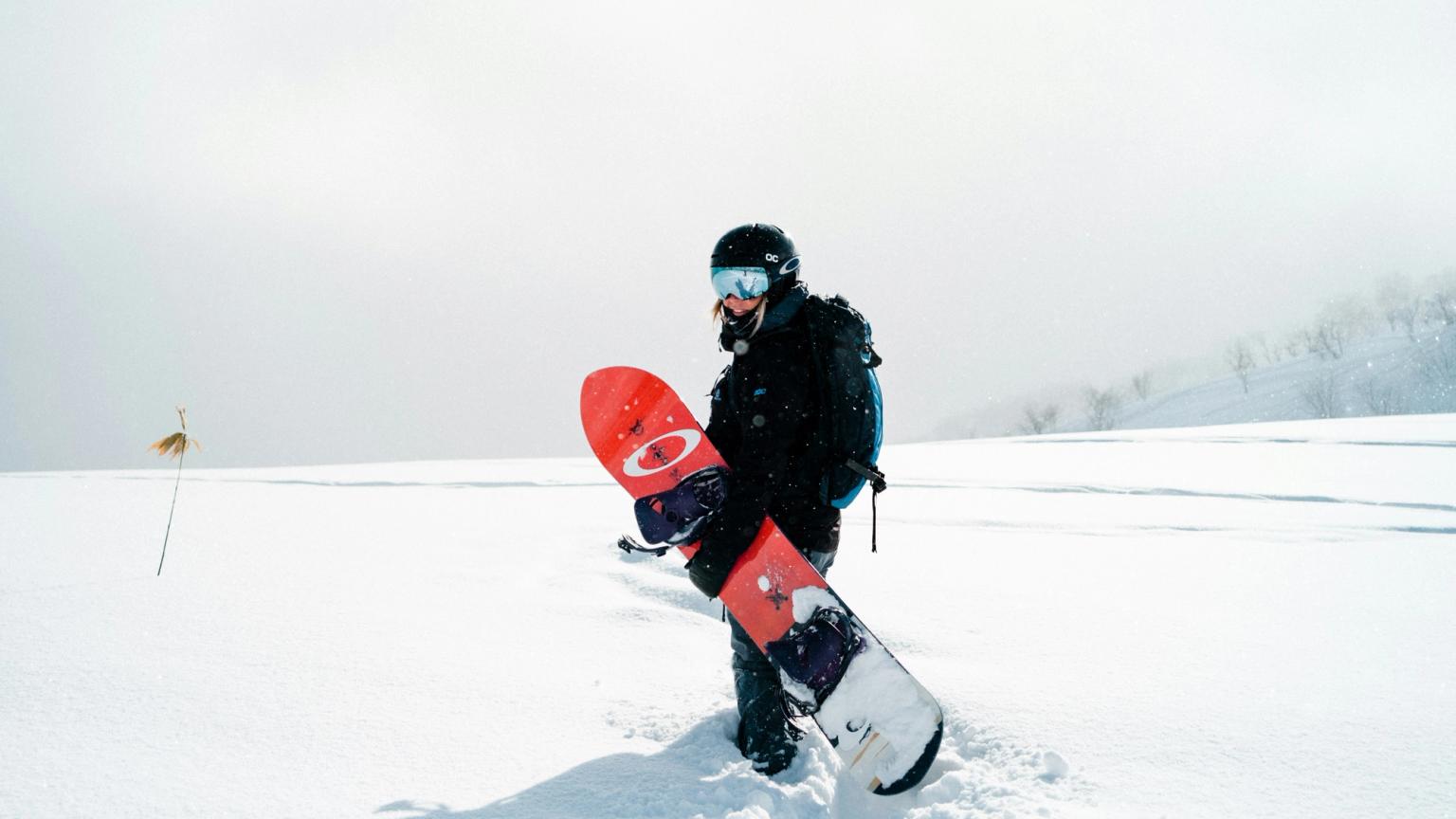 En snowboardkjører med hjelm og skibriller går gjennom dyp snø og bærer et rødt snowboard.
