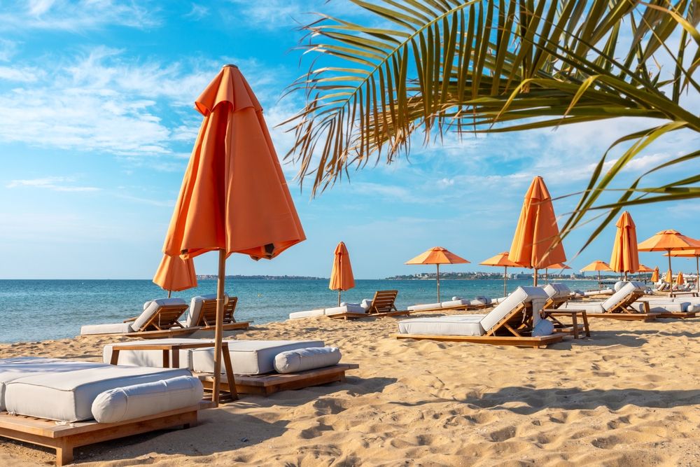 Oransje strandparasoller og solsenger på en sandstrand ved havet under en blå himmel.
