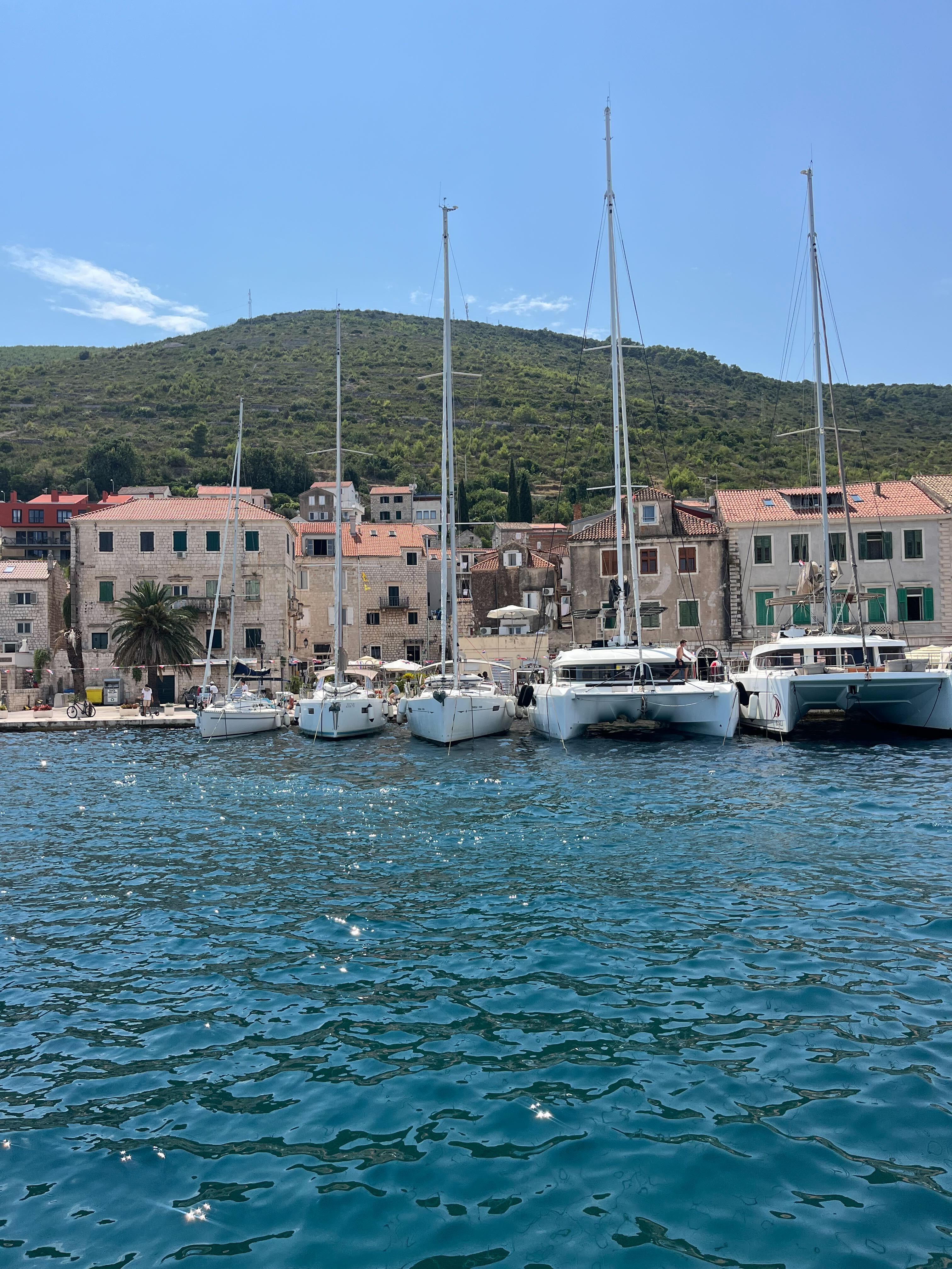 Des bateaux amarrés dans un port ensoleillé en Croatie, devant une ville côtière et un versant de colline verdoyant.