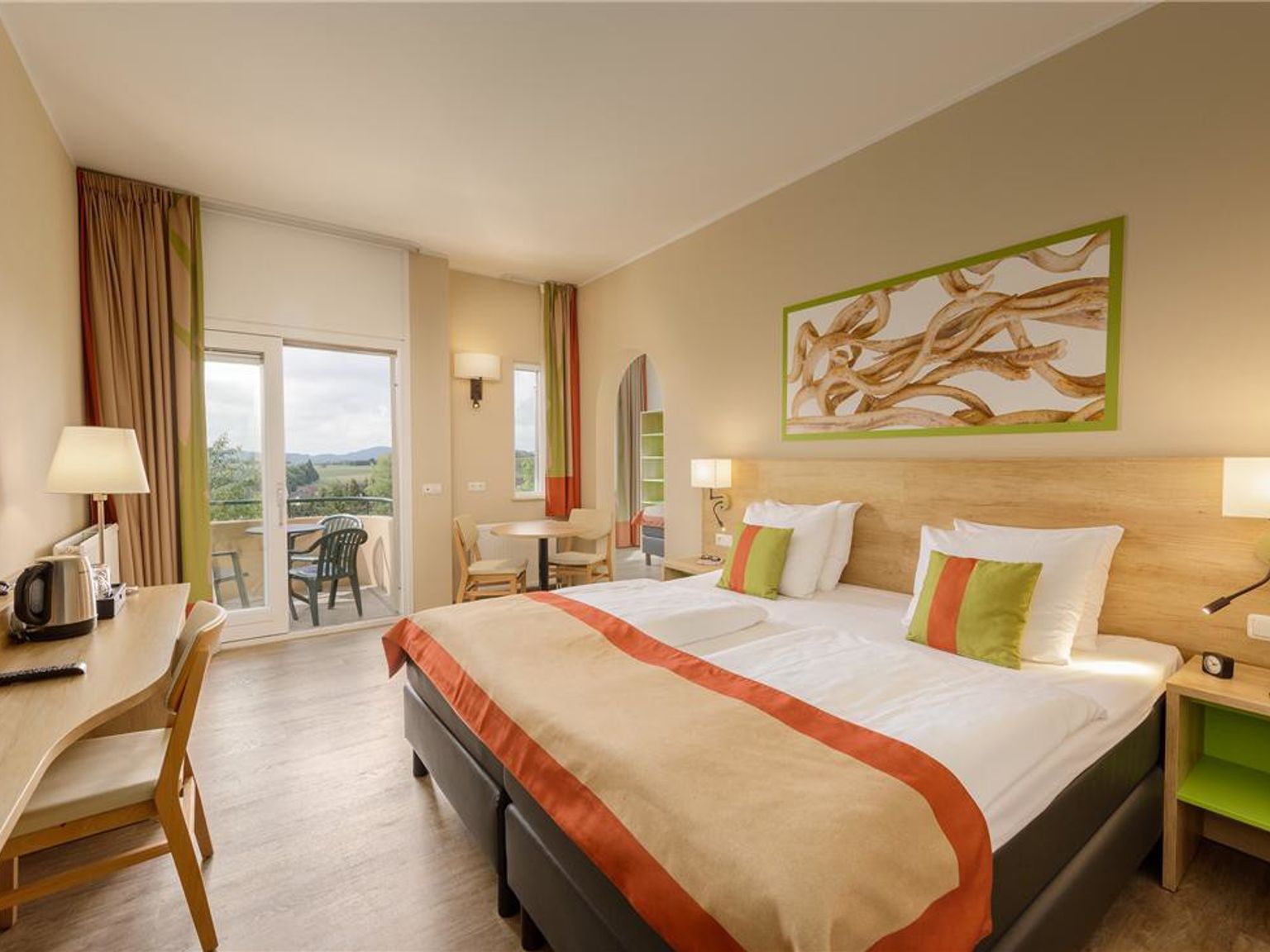 Een lichte hotelkamer met een groot bed, houten bureau en een balkon met uitzicht op groene heuvels. Center Parcs Park Hochsauerland