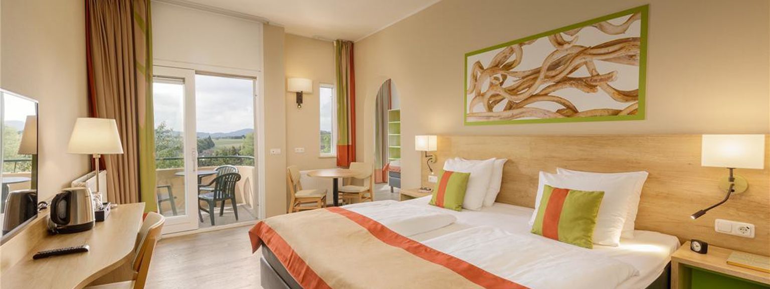 Een lichte hotelkamer met een groot bed, houten bureau en een balkon met uitzicht op groene heuvels. Center Parcs Park Hochsauerland