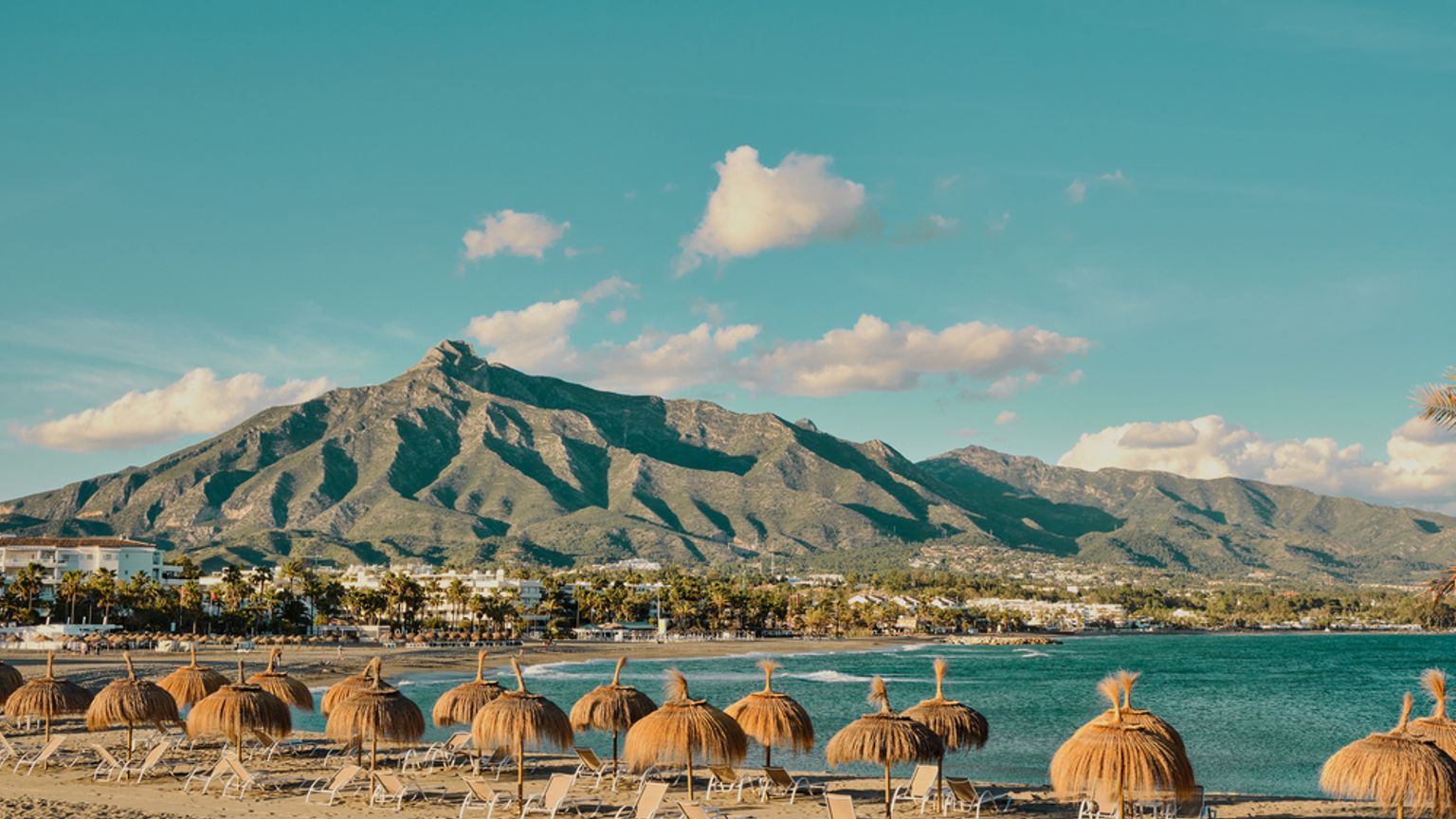 rannalla on paljon sateenvarjoja ja tuoleja, ja taustalla näkyy vuoria.