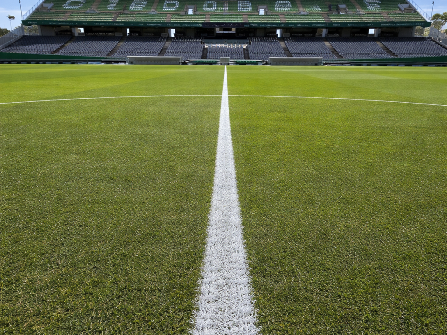 Empty sports stadium with a green field and a white center line leading to spectator stands under a blue sky.