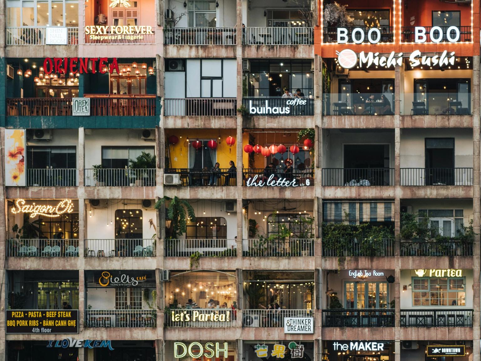 En fler-etagers bygning tæt pakket med caféer, restauranter og butikker, hver enhed med en balkon med et klart oplyst skilt. Ho Chi Minh City