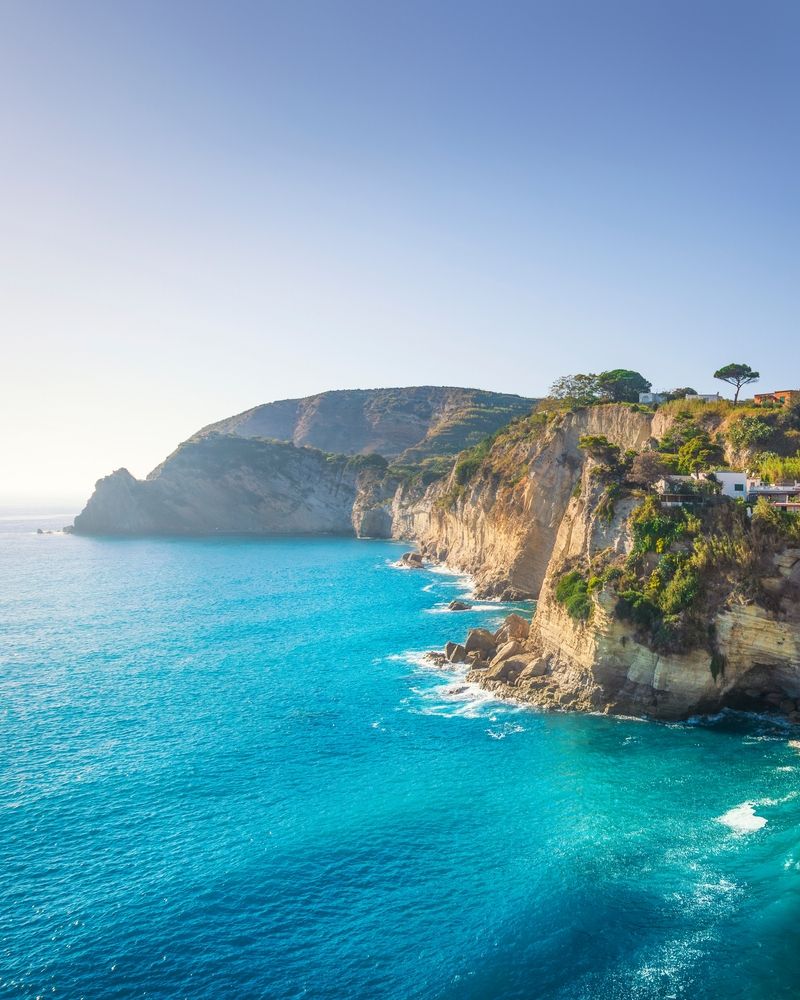 Une mer bleu vif rencontre des falaises rocheuses spectaculaires couvertes de végétation verte, avec quelques bâtiments blancs au sommet. Ischia, Italie
