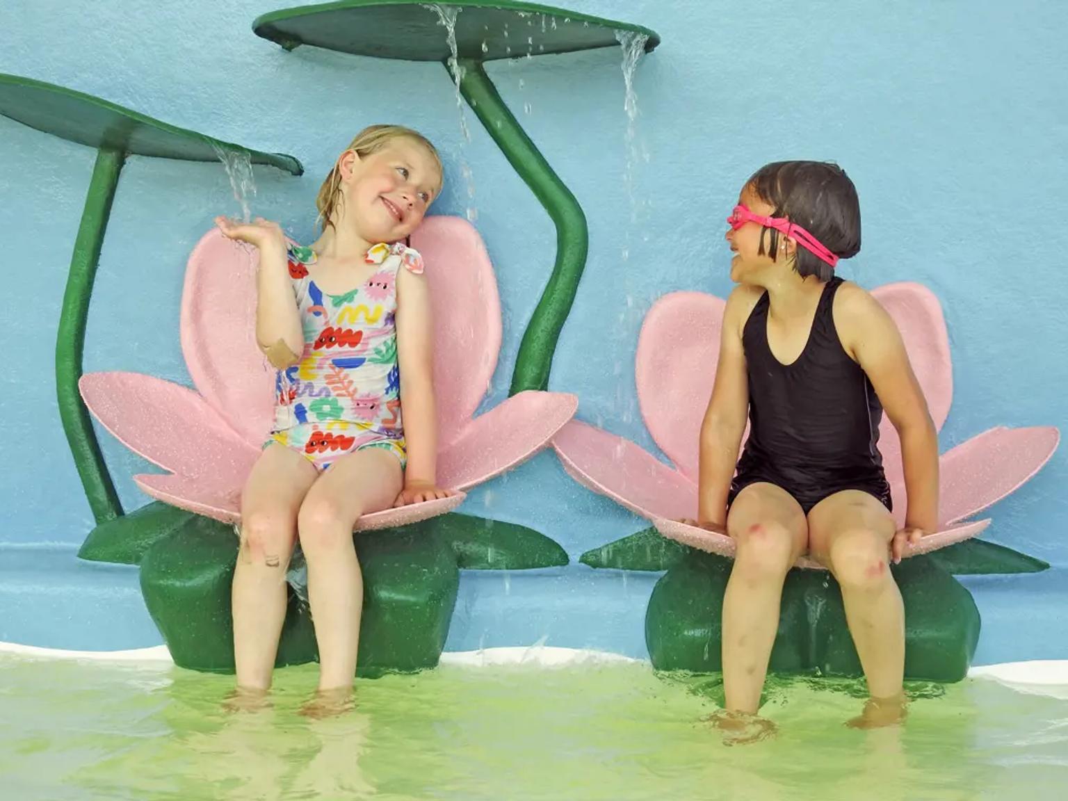 To smilende jenter sitter på lotusformede vannfunksjoner i en vannlekeplass.