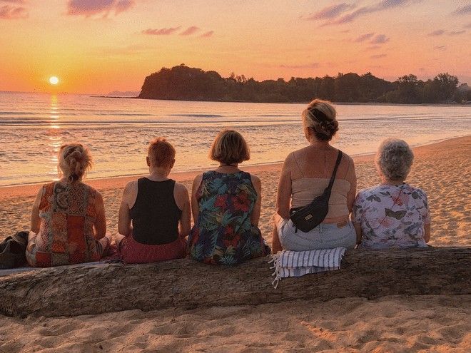 Fem kvinnor sitter på en stock på stranden och ser en ljust orange solnedgång över havet.