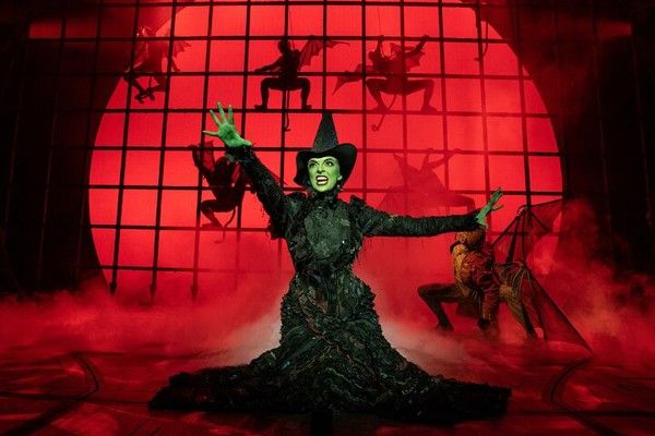 Elphaba, the green-skinned Wicked Witch, in a black dress and hat, poses dramatically on a red-lit stage with flying monkeys.