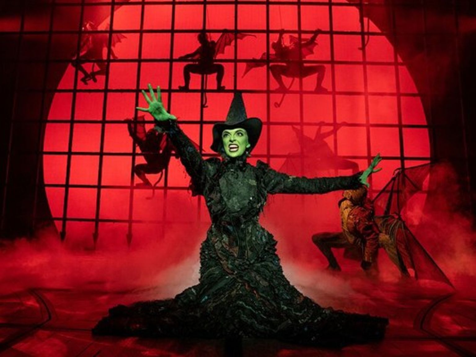 Elphaba, the green-skinned Wicked Witch, in a black dress and hat, poses dramatically on a red-lit stage with flying monkeys.