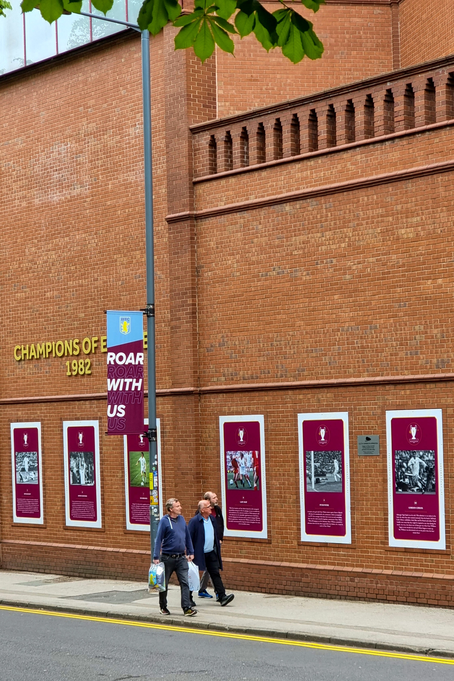 A brick building with "CHAMPIONS OF EUROPE 1982" in gold lettering and framed historical football images on the wall. Three men walk on the sidewalk below.