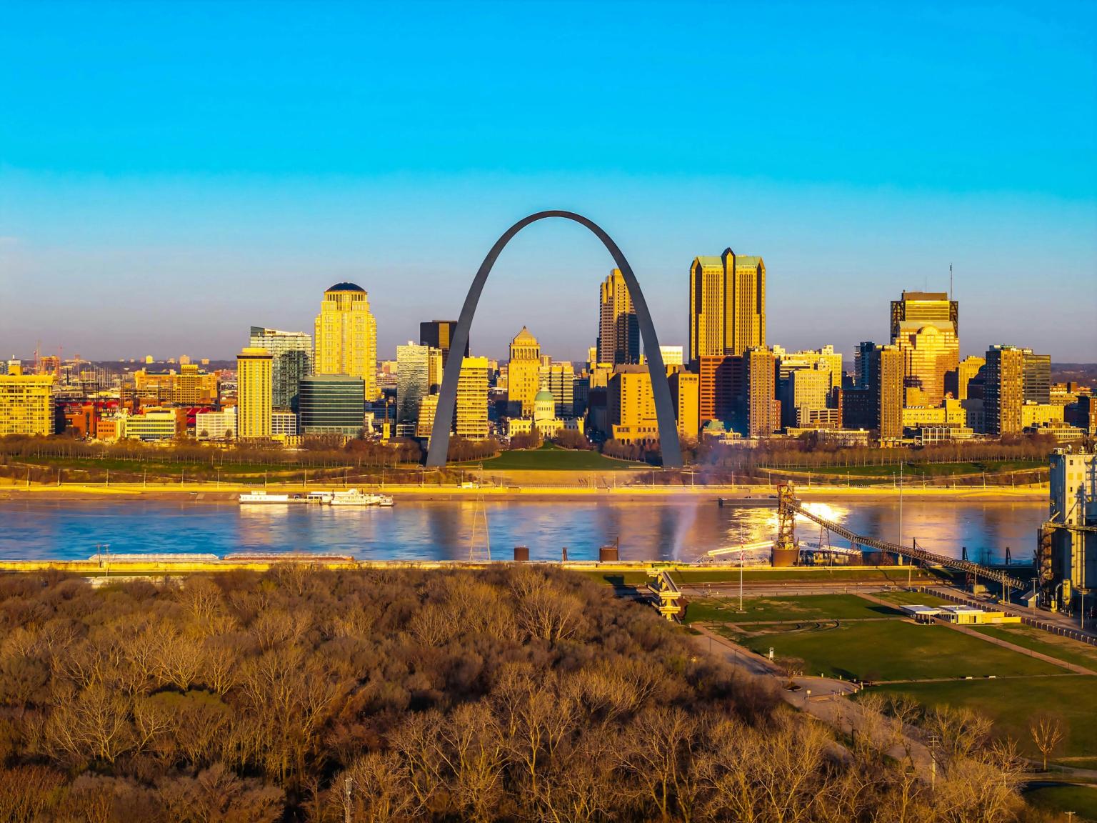 Bybillede af St. Louis med Gateway Arch, Mississippi-floden og nøgne træer i varmt, gyldent lys.