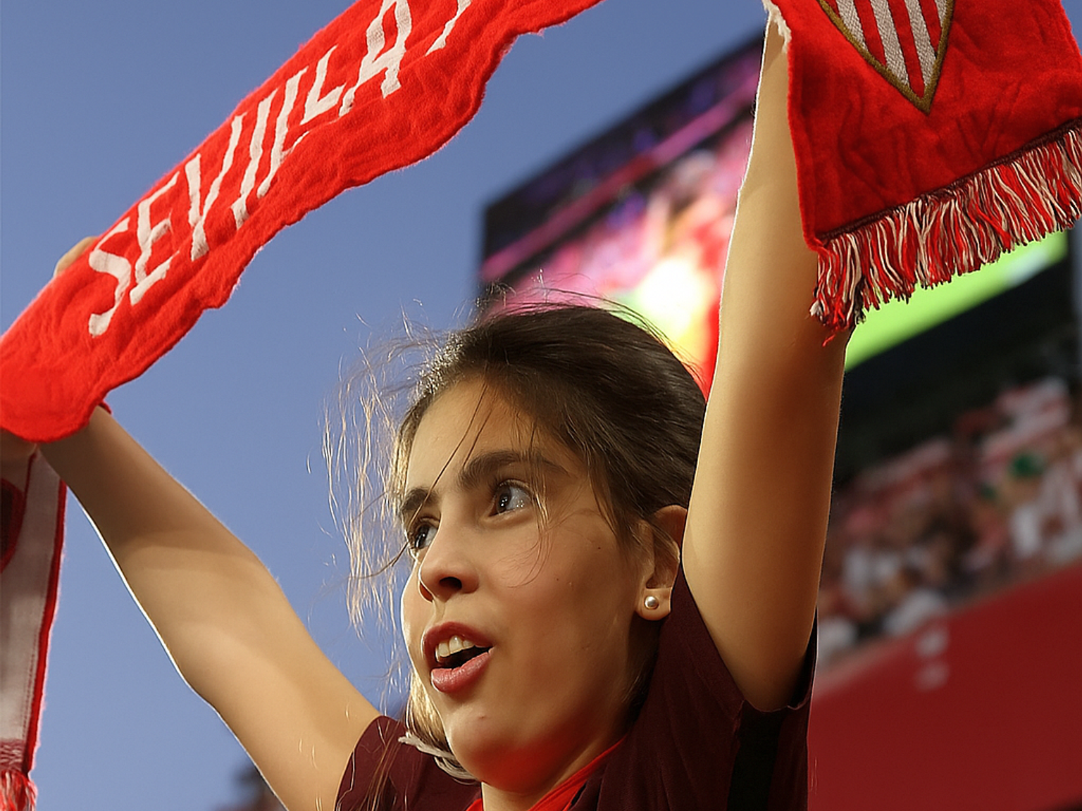 En ung fan i en rød Sevilla FC-trøye holder et rødt "Sevilla FC"-skjerf over hodet og heier på en fotballstadion.