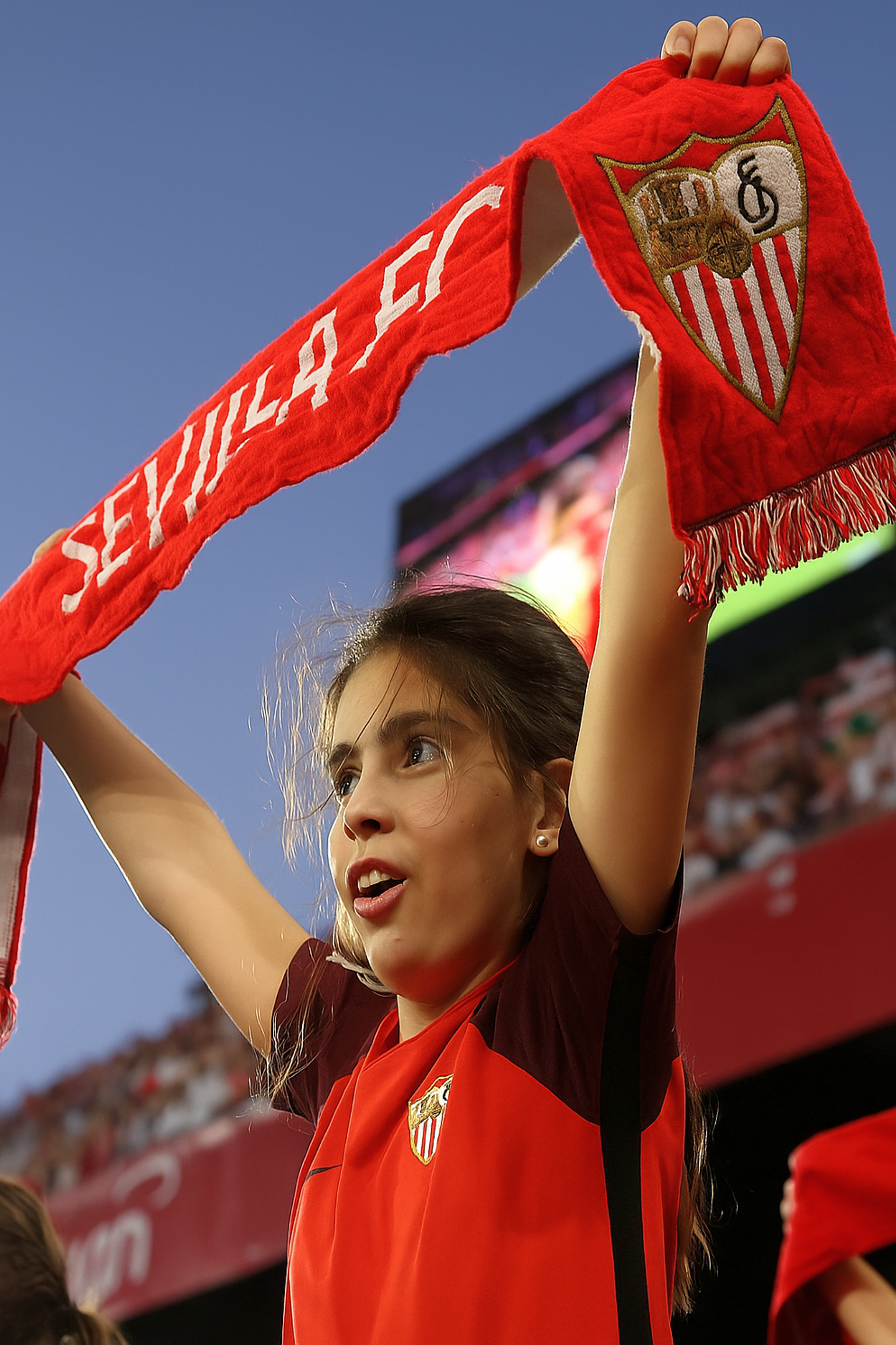 En ung fan i en rød Sevilla FC-trøye holder et rødt "Sevilla FC"-skjerf over hodet og heier på en fotballstadion.