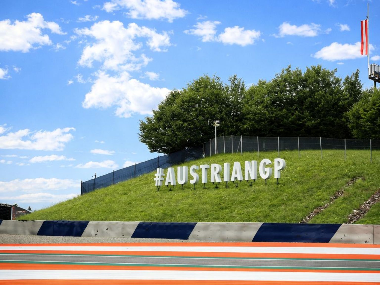 A black and yellow formula racing car on a track with a large "#AUSTRIANGP" sign on a grassy hill under a blue sky.