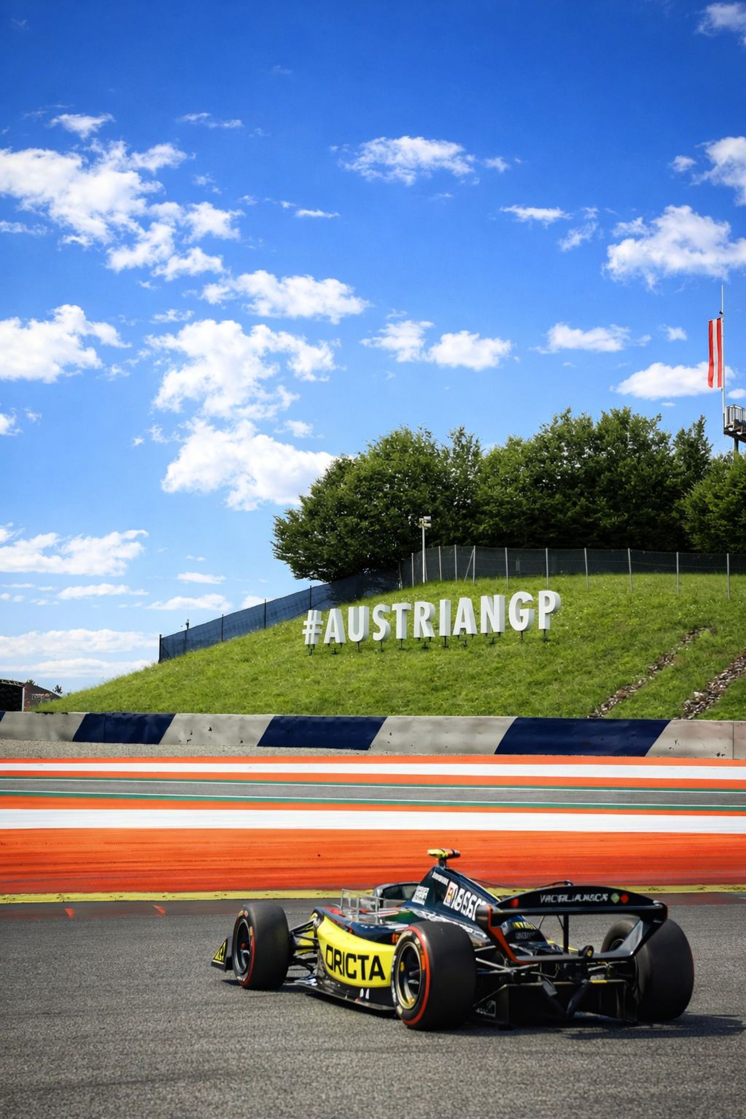 A black and yellow formula racing car on a track with a large "#AUSTRIANGP" sign on a grassy hill under a blue sky.
