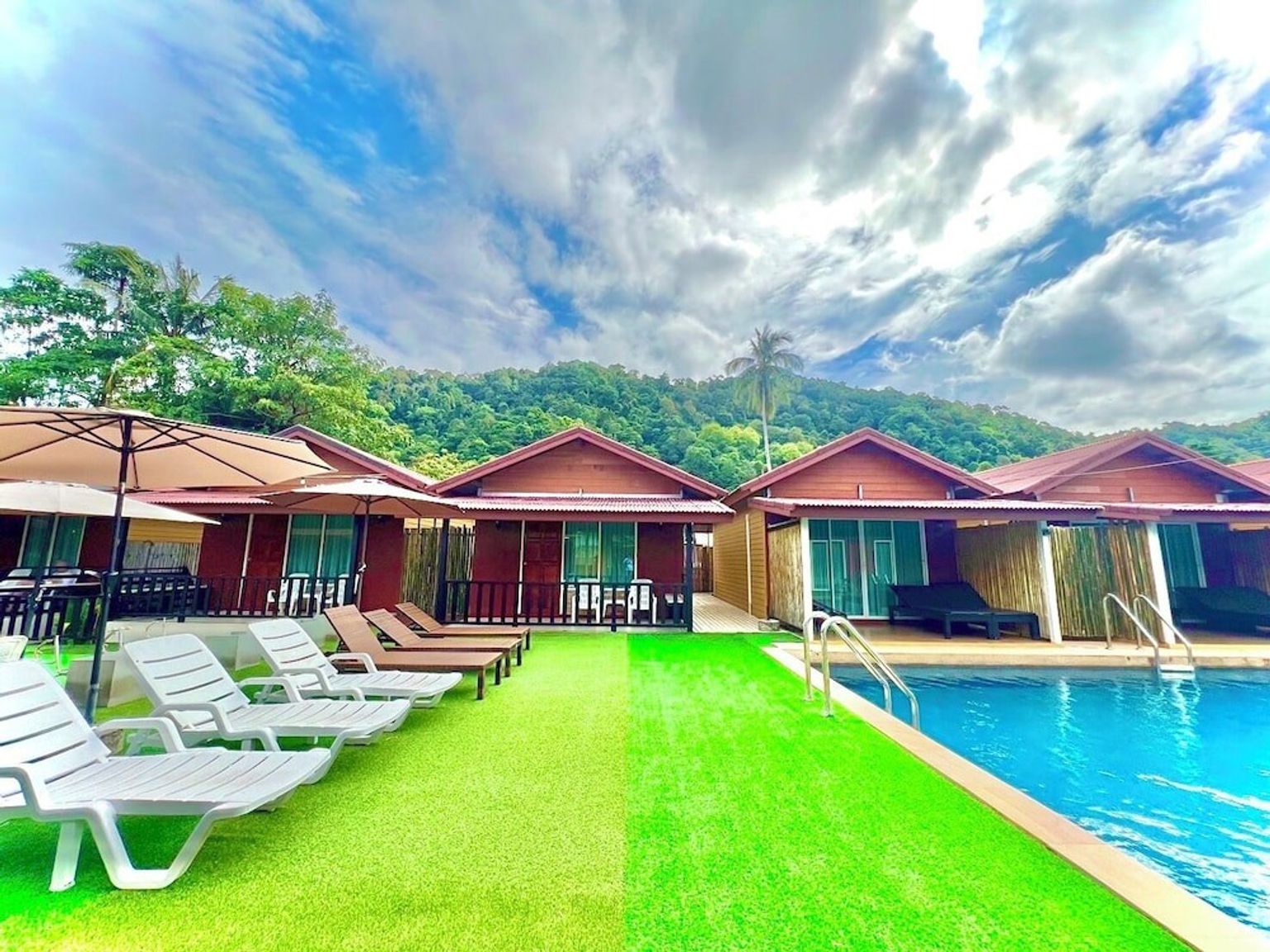 En rad semesterbungalower med solstolar och en pool, med gröna berg i bakgrunden under en delvis molnig himmel.