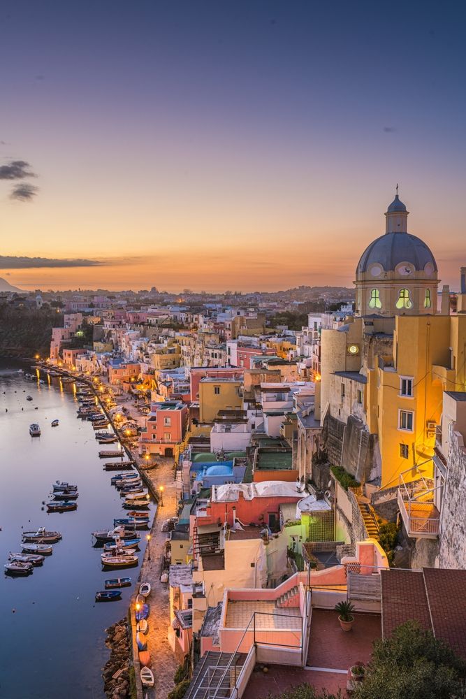 Ville côtière animée au crépuscule avec des bâtiments aux couleurs vives, une église à dôme et de nombreux bateaux le long d’un port illuminé. Procida, Italie