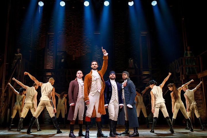 Holdet fra Hamilton optræder på scenen, med hovedrollerne i koloniale kostumer og ensemblets dansere i træningstøj, dramatisk oplyst af spotlights.