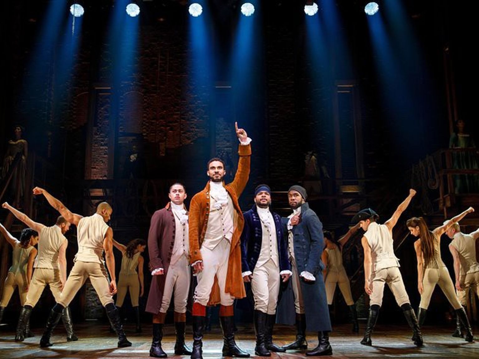 Holdet fra Hamilton optræder på scenen, med hovedrollerne i koloniale kostumer og ensemblets dansere i træningstøj, dramatisk oplyst af spotlights.